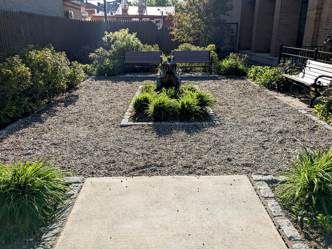 A perspective view of a park area from the entrance with benches, bushes, a center sculpture, and a crushed stone walkway.