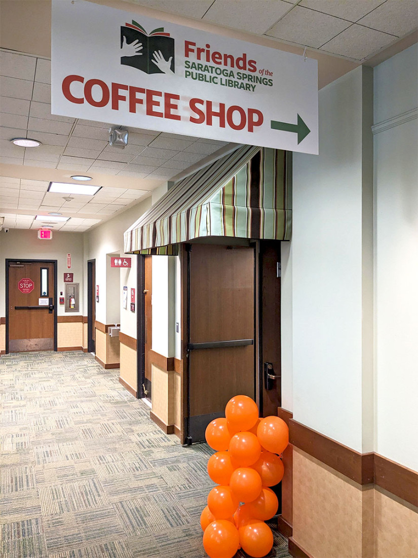 A “Coffee Shop” sign hangs in front of an awning to doors, with orange celebratory balloons to grab attention.