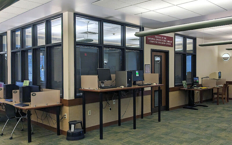 A doorway surrounded by 3 computers along a wall. Lights are on inside the room, with closed metal shades over the windows.