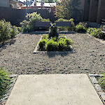 Thumbnail: A perspective view of a park area from the entrance with benches, bushes, a center sculpture, and a crushed stone walkway.