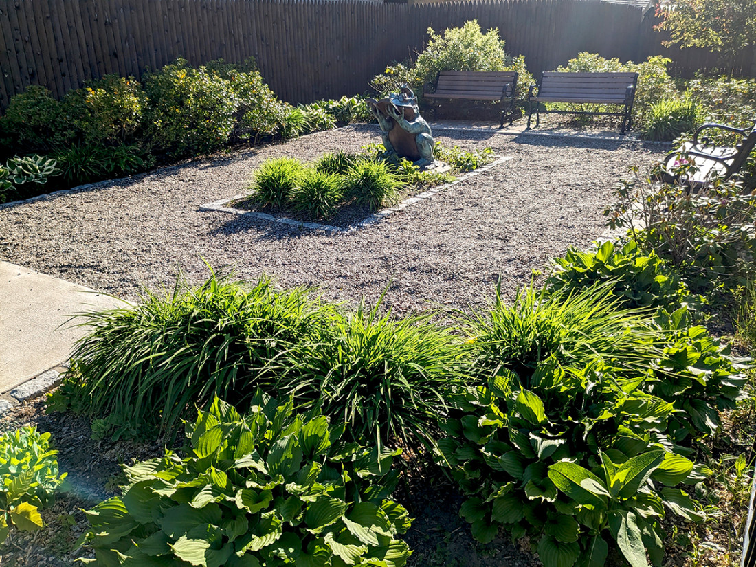 A perspective view of a park area from the right-side of the entrance with benches, bushes, a center sculpture, and a crushed stone walkway.