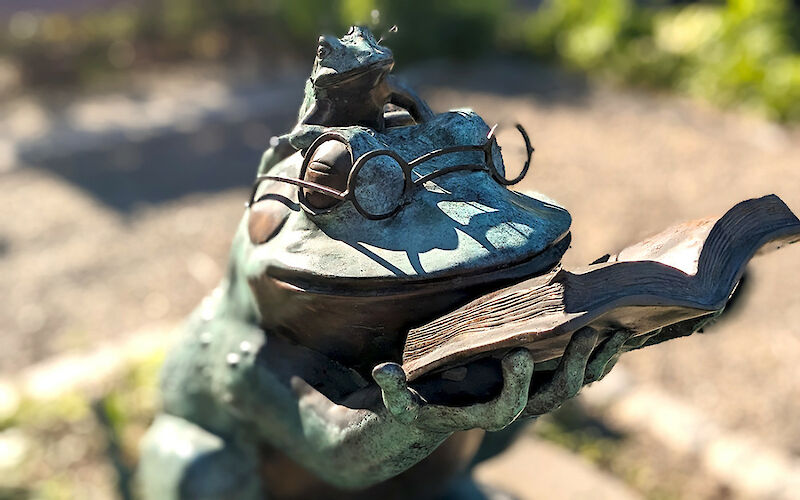 A sculpture of a large from reading a story from a book to a smaller frog that is sitting atop its head.