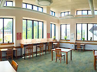 Tables with chairs around a room’s perimeter, against the walls. One lone table and chair in the center of the floor. Tall windows cover the majority of the walls.