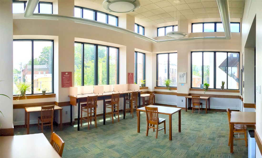 Tables with chairs around a room’s perimeter, against the walls. One lone table and chair in the center of the floor. Tall windows cover the majority of the walls.