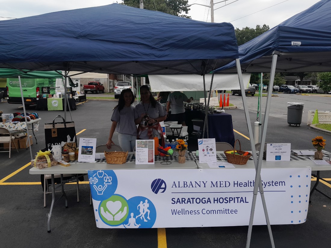 Two staff members from Saratoga Hospital under a blue tent standing behind a table with information and a white Saratoga Hospital banner hanging from front of table