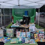 Thumbnail: A panel van, decorated with the library’s colors of green and white.  Picture books display on a table with a black tablecloth and green runner.  White tent overhead.  Rain in the background.