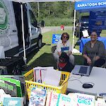 Thumbnail: Two people. one from the library, and a therapy dog sitting behind an outdoor book display at a town event