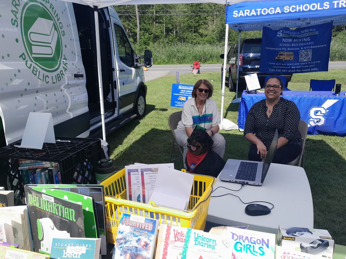 Two people. one from the library, and a therapy dog sitting behind an outdoor book display at a town event