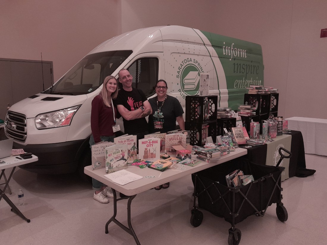 a panel van with 3 people in front of van. Table with books in front of 3 people and van