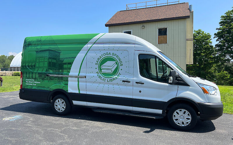 A panel van, decorated with the library’s colors of green and white showcasing a two-tone photo depicting the library all in green on the back half and the library logo in green on white on the front.