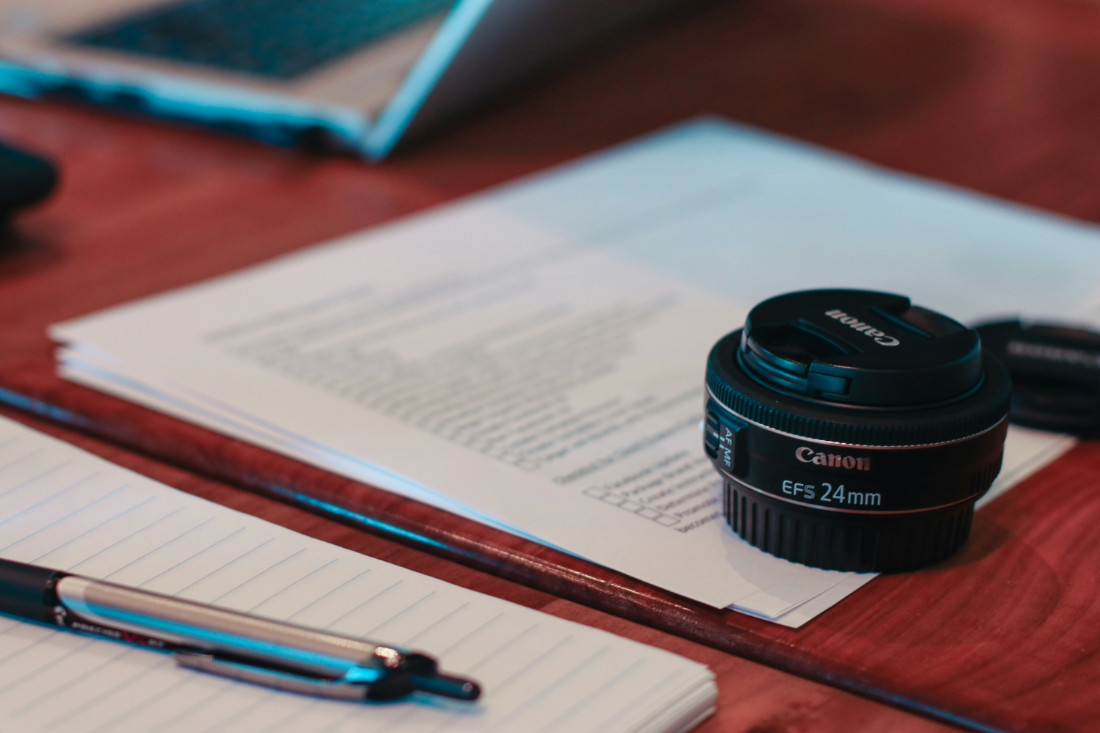 A wooden desk with printed paper slightly out of focus, and in the foreground a pen and camera lens.