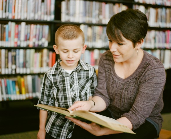 A woman and child standing in front of book shelves, kneels down and holds a book open and points to a section, as if reading, to the child.